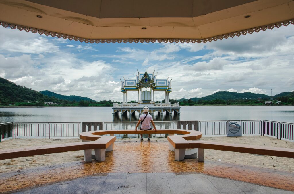 A person sitting on a bench overlooking a tranquil lake and ornate pavilion under cloudy skies.
